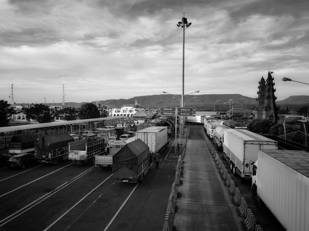 Black and white image of a bustling shipping port with trucks and ferries under a cloudy sky.